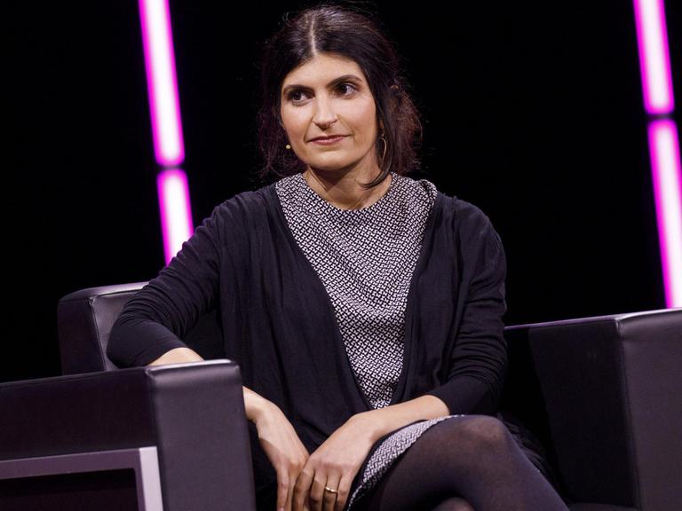 Shida Bazyar auf einem Sessel auf einer Bühne, im Hintergrund sind rosa Neonröhren zu sehen