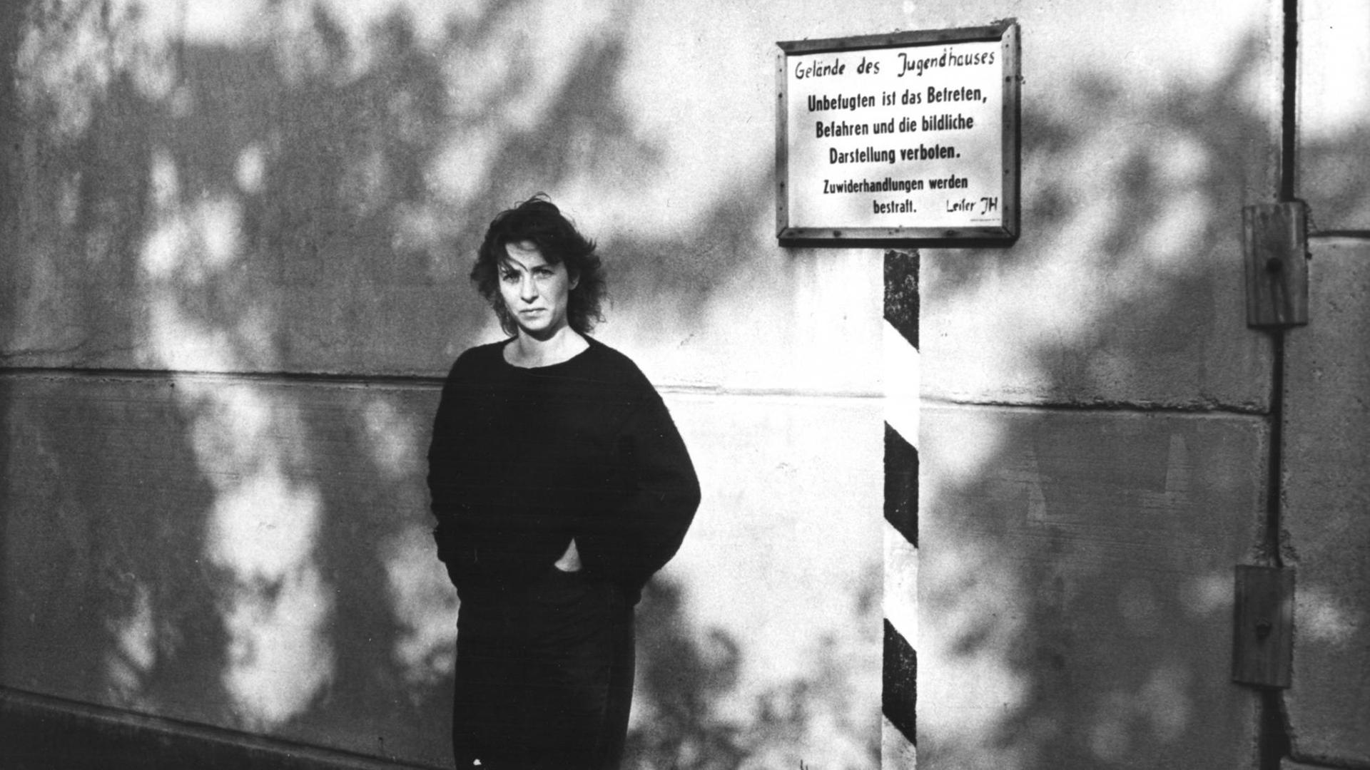 Die Regisseurin Sibylle Schönemann steht in ihrem Film "Verriegelte Zeit" vor einer Außenwand. Auf einem an der Wand angebrachten Schild steht geschrieben: "Geländes des Jugendhauses. Unbefugten ist das Betreten, Befahren und die bildliche Darstellung verboten, Zuwiderhandlungen werden bestraft. Leiter JH."