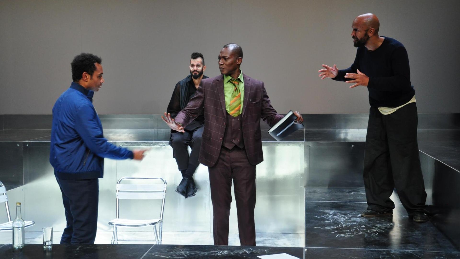 Vier schwarze Männer auf der Bühne. Drei sprechen im Stehen miteinander, einer sitzt auf einem Stuhl im Hintergrund. Eine Szene aus dem Theaterstück: "Walking large" von Toks Körner