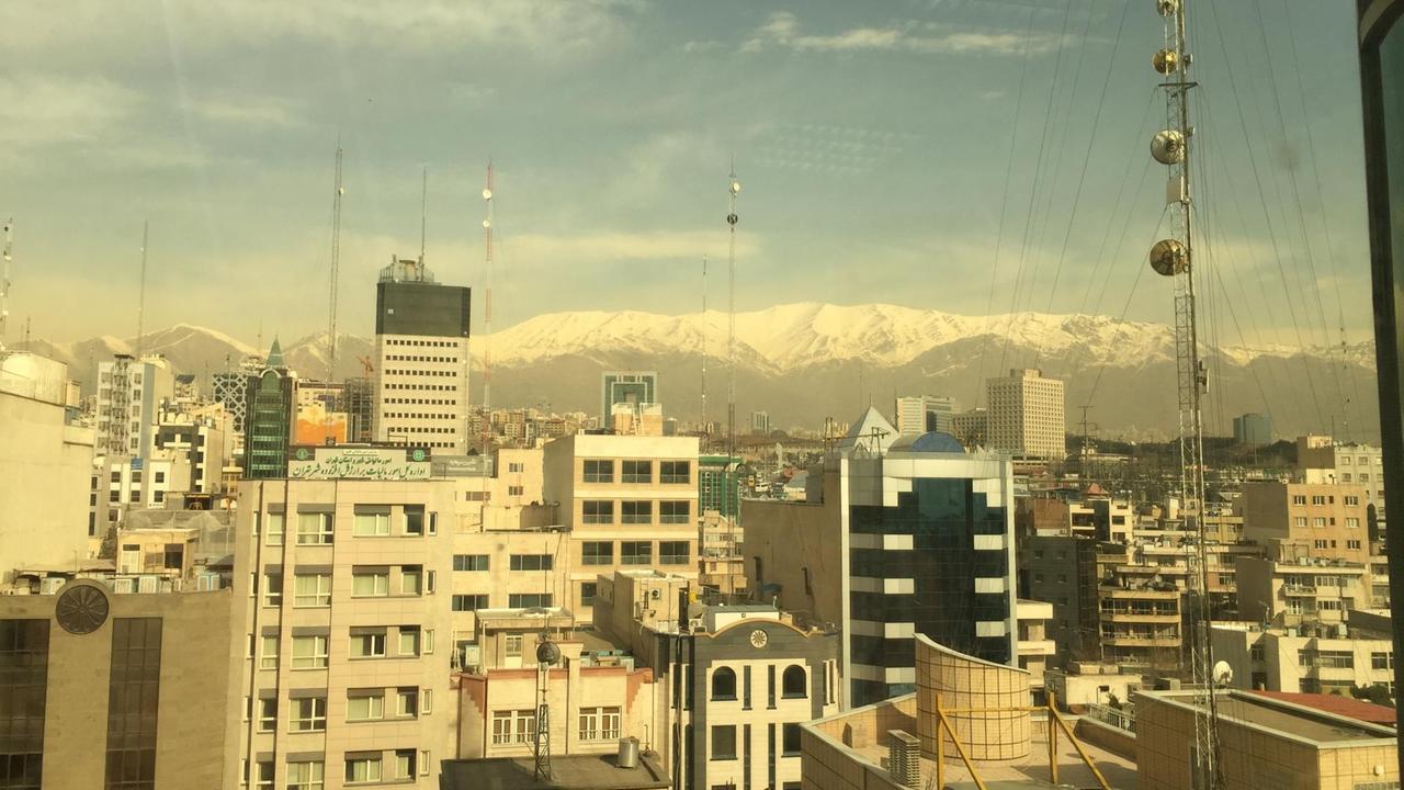 Blick auf Wohnhäuser und Wolkenkratzer der iranischen Hauptstadt, im Hintergrund verschneite Berggipfel unter blauem Himmel.