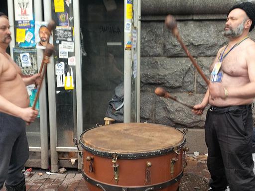 Zwei Männer mit nackten Oberkörpern stehen sich auf einer Straße vor einem Gebäude gegenüber und trommeln auf großen Standtrommeln.