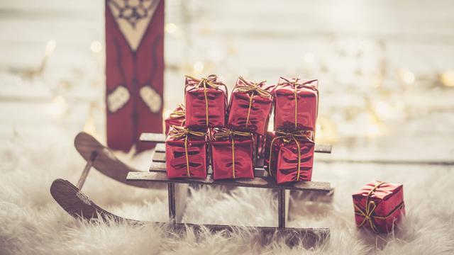 Eine kleine Weihnachtsmannfigur steht hinter einem Holzschlitten, der mit roten Geschenken beladen ist.