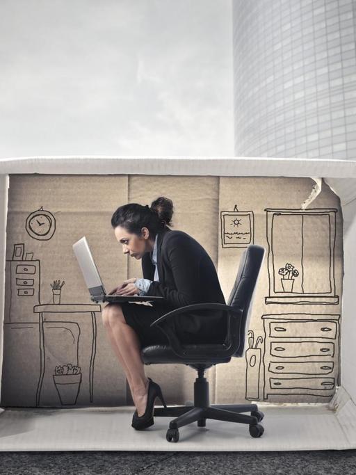 Illustration (Collage) einer Business-Frau, die in einem Karton-Büro sitzt, umgeben von einer grauen Hochhauswelt