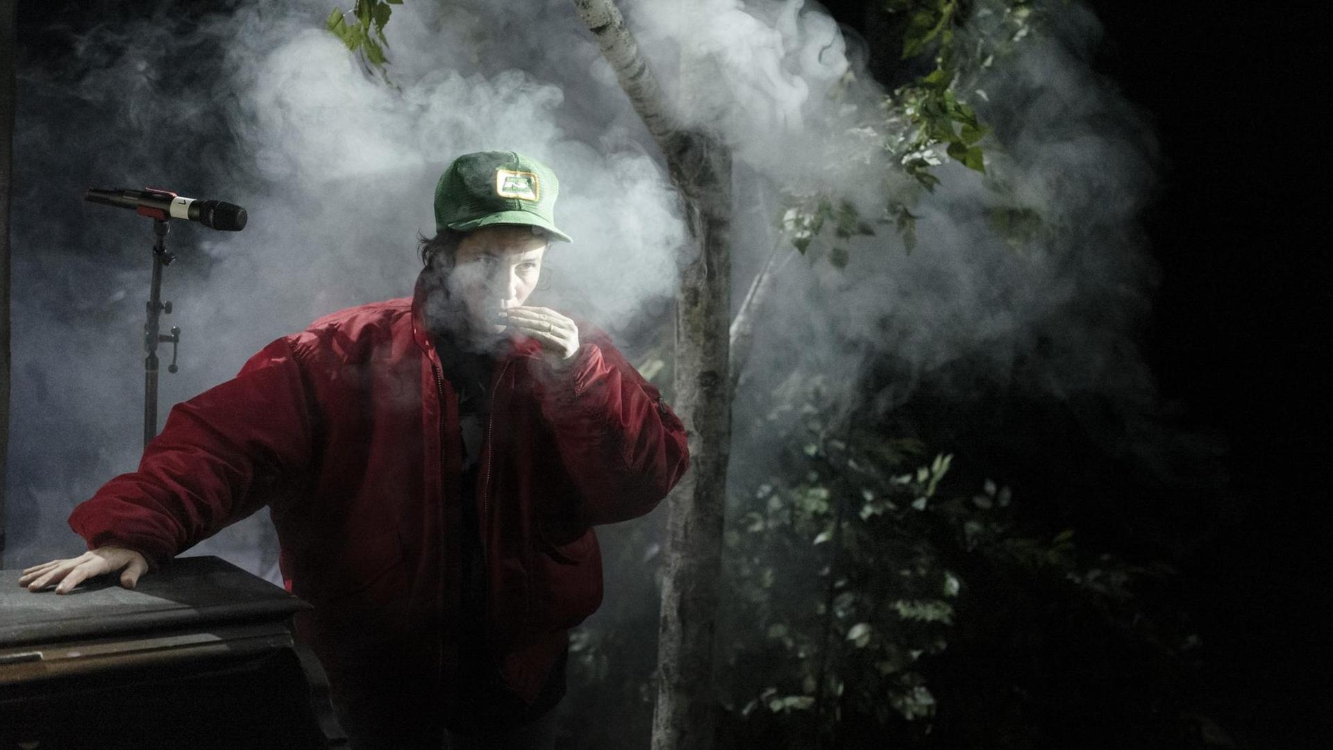 Ein junger Mann mit Basekap raucht im Wald, an ein Klavier gelehnt, düstere Bildstimmung.