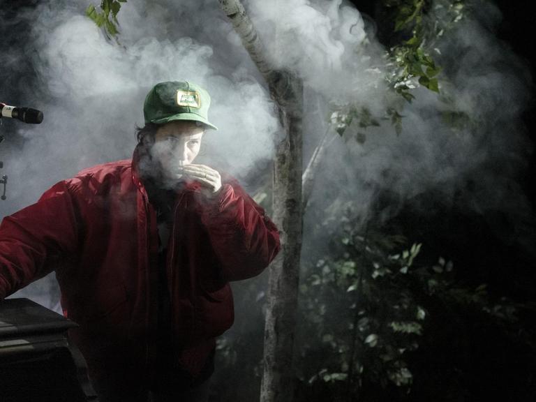 Ein junger Mann mit Basekap raucht im Wald, an ein Klavier gelehnt, düstere Bildstimmung.