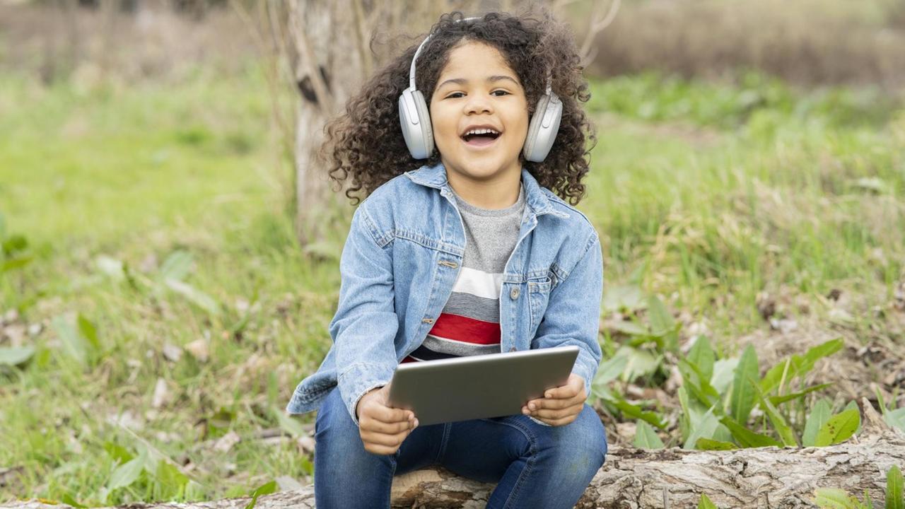 Junge sitzt mit I-Pad und Kopfhörer im Grünen Junge sitzt mit I-Pad und Kopfhörer im Grünen
