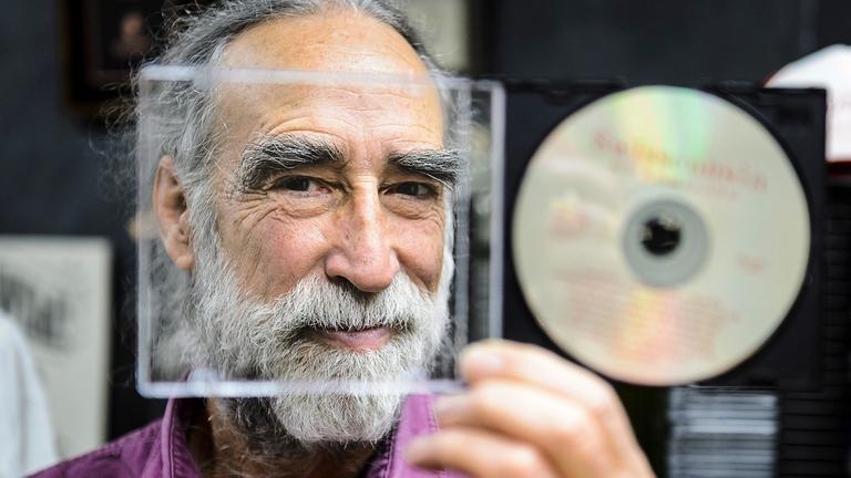 Ein Mann blickt durch ein geöffnetes, leeres, durchsichtiges CD-Cover und lacht.