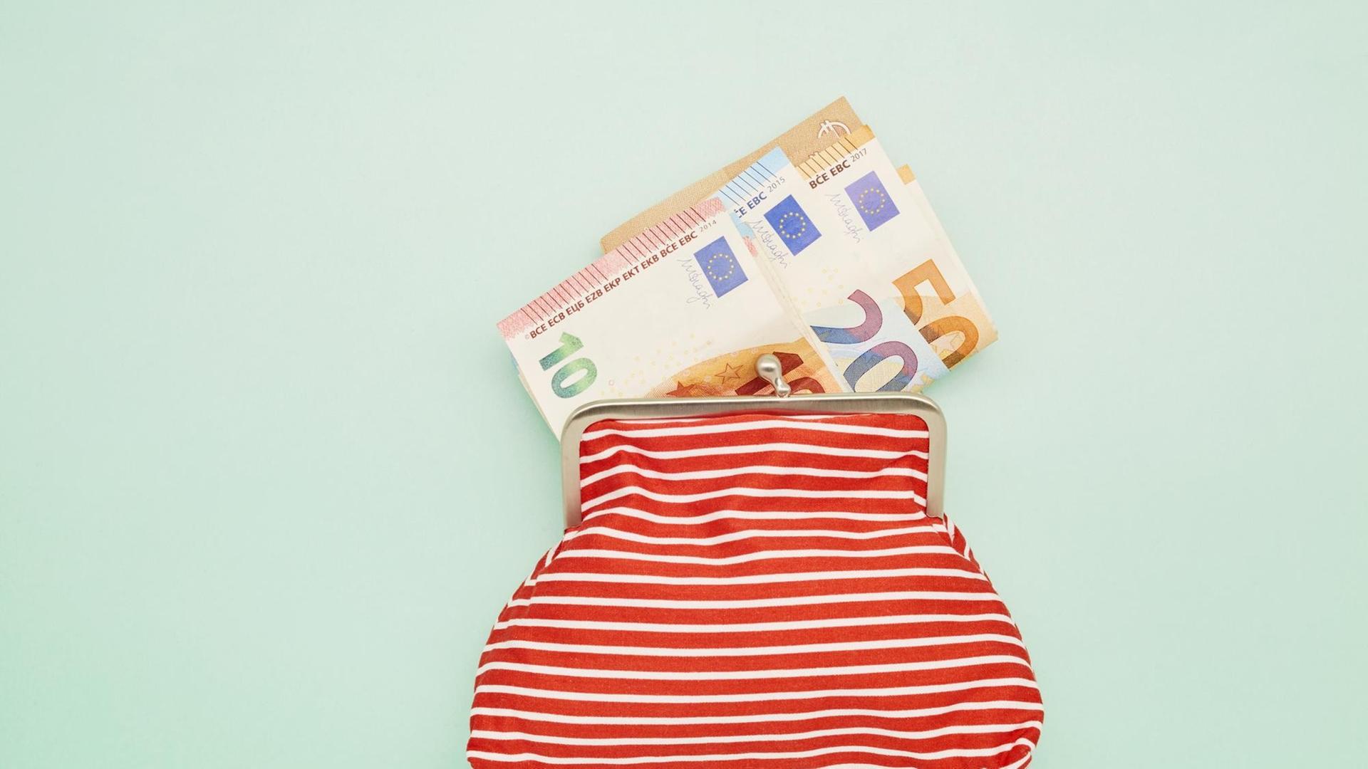 Eine rotweiss gestreifte Geldbörse liegt auf einem mintfarbenen Untergrund. Daraus schauen einige Euronoten. Eine rotweiss gestreifte Geldbörse liegt auf einem mintfarbenen Untergrund. Daraus schauen einige Euronoten.