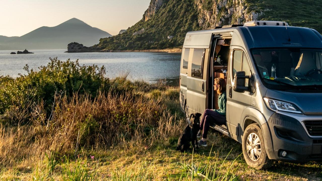 Eine Frau sitzt in der geöffneten Schiebetür ihres Camper-Vans. Im Hintergrund liegt ein See und ein Berg. Eine Frau sitzt in der geöffneten Schiebetür ihres Camper-Vans. Im Hintergrund liegt ein See und ein Berg.