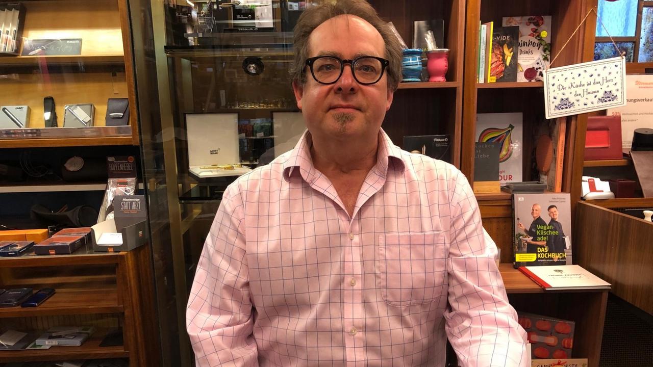 Porträt. Martin Herrmann sitzt im rosa Hemd hinter dem Ladentisch. Hinter ihm ein Regal mit Schreibutensilien. Porträt. Martin Herrmann sitzt im rosa Hemd hinter dem Ladentisch. Hinter ihm ein Regal mit Schreibutensilien.