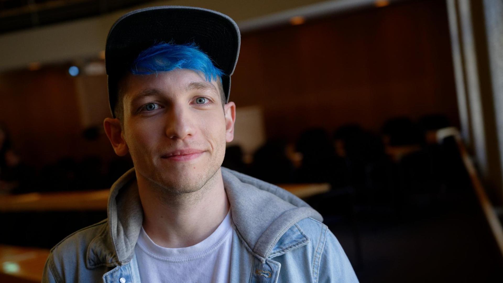 EIn junger Mann mit blauen Haaren, er trägt Jeansjacke, T-Shirt und Käppi, in einem Veranstaltungssaal. Es handelt sich um den Youtuber Rezo. EIn junger Mann mit blauen Haaren, er trägt Jeansjacke, T-Shirt und Käppi, in einem Veranstaltungssaal. Es handelt sich um den Youtuber Rezo.