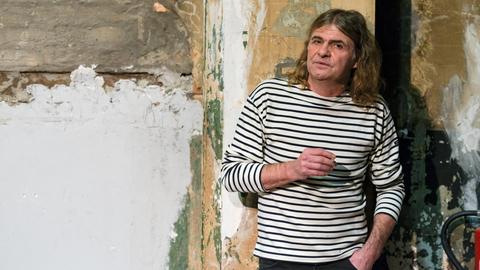 Der Musiker Wenzel mit Zigarette in der Hand vor einer heruntergekommenen Wand