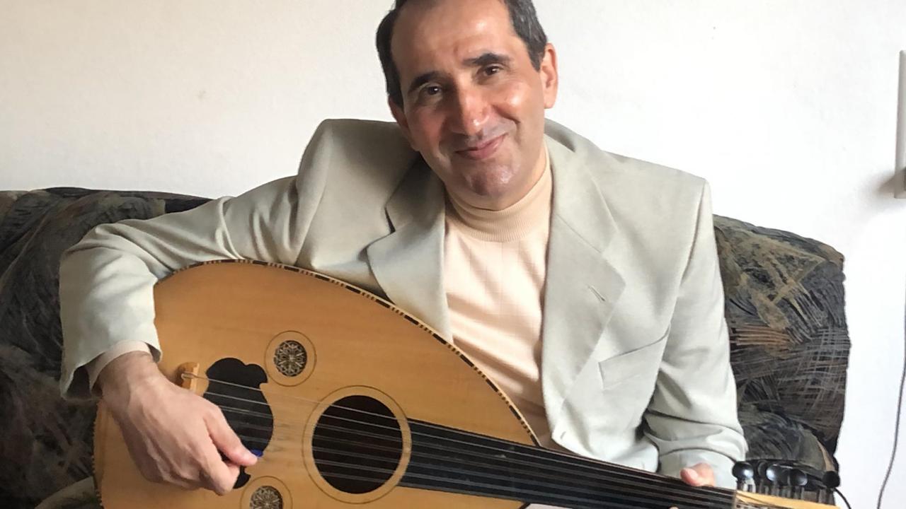 Ein lächelner Mann mit kurzen dunklen Haaren und einer Oud (arabisches Instrument) auf dem Schoß.