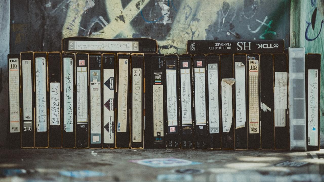Eine Reihe VHS-Kassetten stehen vor einer mit Graffiti bemalten Wand