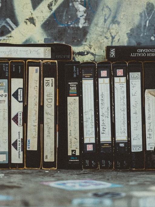 Eine Reihe VHS-Kassetten stehen vor einer mit Graffiti bemalten Wand