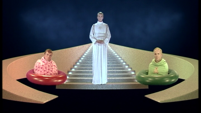 Videostill vom Künstler Bjørn Melhus, eine priesterliche Figur steht in der Mitte auf einer Treppe, daneben zwei Menschen in Schwimmreifen