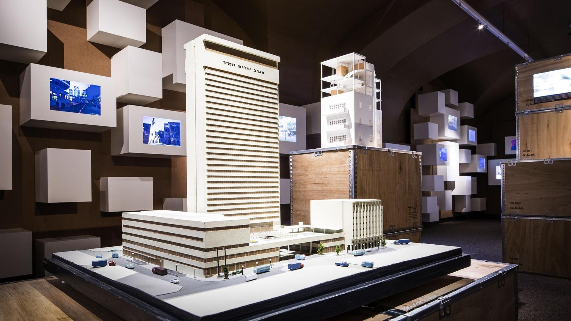 Innerhalb der Ausstellung stehen Archtitekturmodelle auf hölzernen Transportkisten zwischen Videoprojektionen Innerhalb der Ausstellung stehen Archtitekturmodelle auf hölzernen Transportkisten zwischen Videoprojektionen