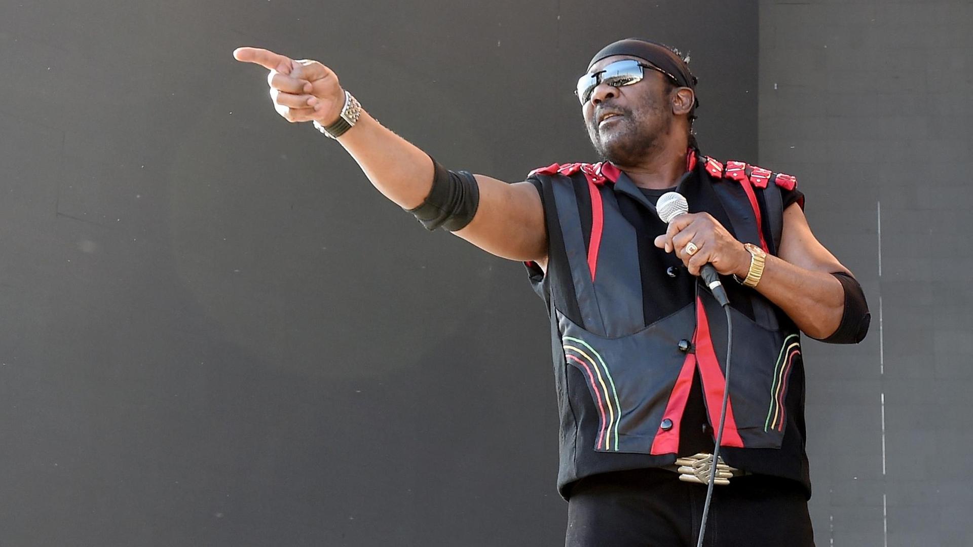 Ein Mann mit Sonnenbrille vor den Augen steht auf einer Bühne und hält in der linken Hand ein Mikrofon. Mit der Rechten streckt er den Zeigefinger am waagerecht gehobenen Arm.