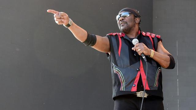 Ein Mann mit Sonnenbrille vor den Augen steht auf einer Bühne und hält in der linken Hand ein Mikrofon. Mit der Rechten streckt er den Zeigefinger am waagerecht gehobenen Arm.