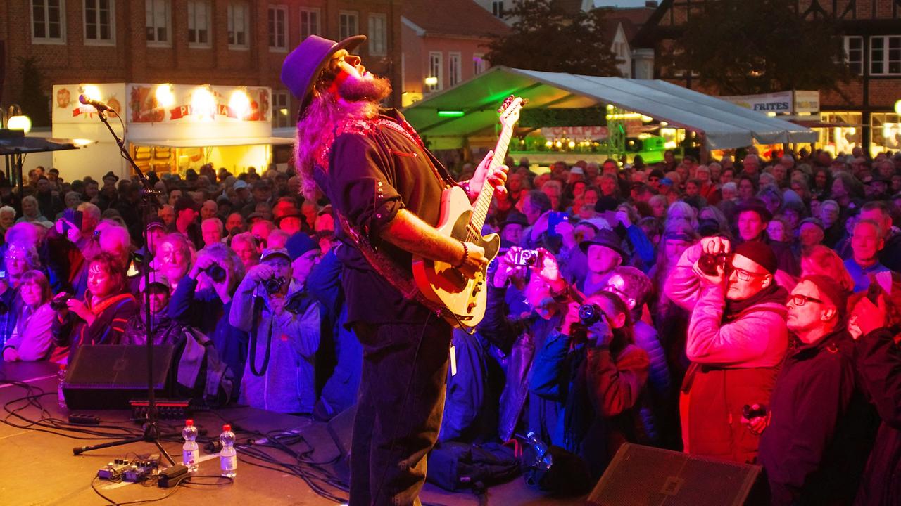 Ein Mann mit Gitarre steht vor Publikum auf einer Bühne. Ein Mann mit Gitarre steht vor Publikum auf einer Bühne.