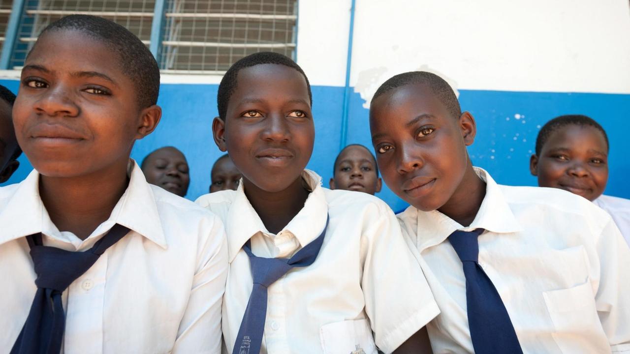 Portrait von drei Mädchen im Teenageralter in einer Schule in Tansania. Sie tragen Schuluniform, weiße Blusen und blaue Krawatten, und lächeln in die Kamera.