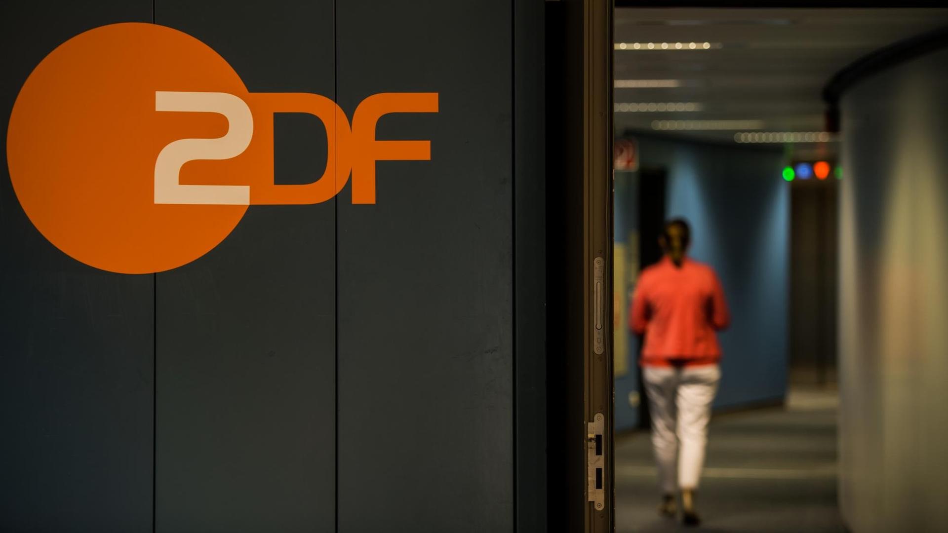 Eine Frau, die von hinten zu sehen ist, läuft durch einen Flur. An einer Wand prangt das orangefarbene Logo des ZDF.