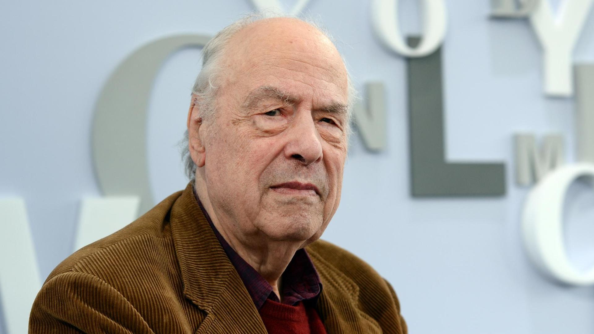 Der 84-jährige Autor Karl-Heinz Bohrer blickt in die Kamera. Er trägt ein braunes Cordsakko und im Hintergrund ist eine Buchstaben-Dekoration der Leipziger Buchmesse zu sehen.