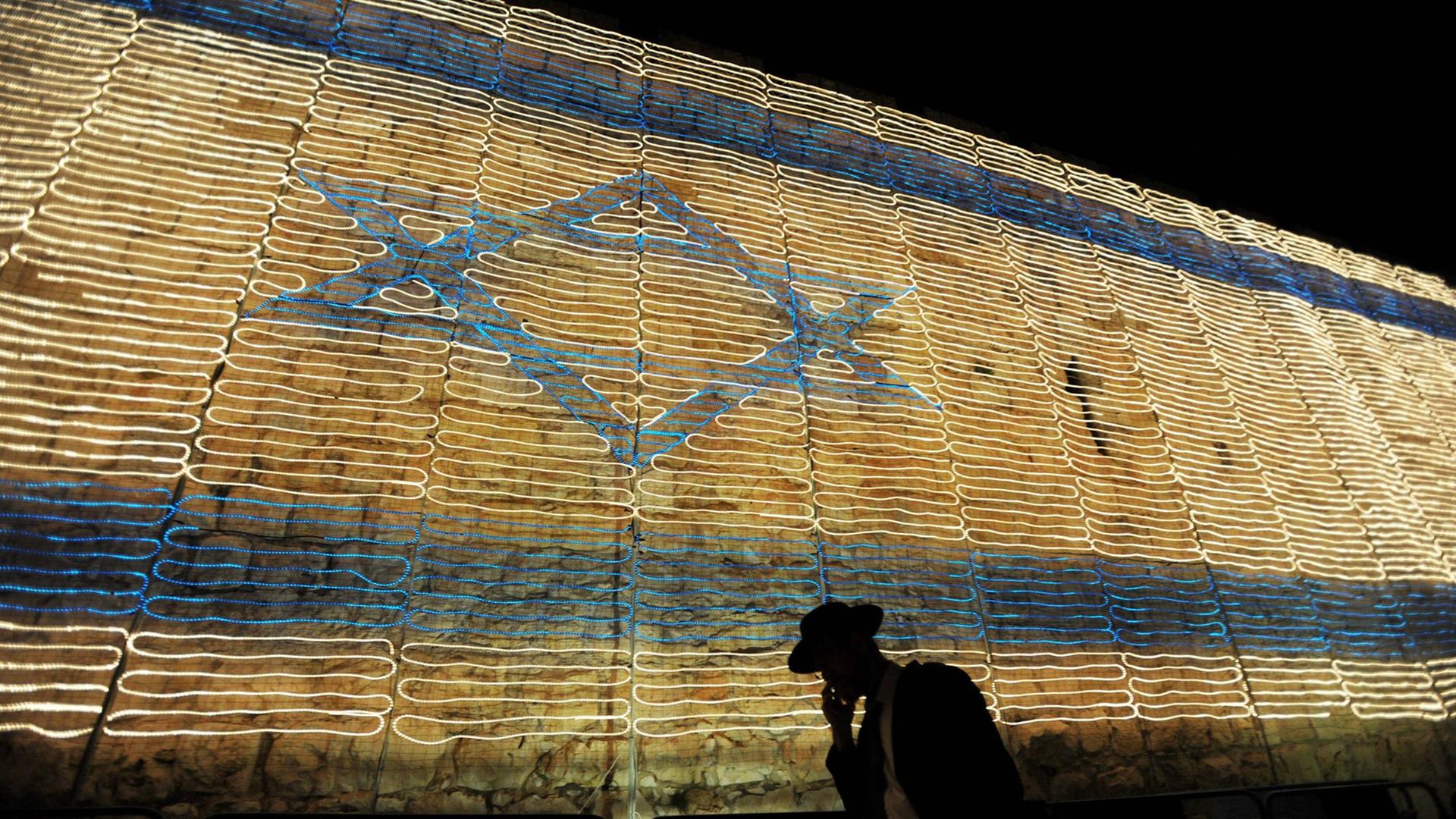 Es ist der 9. Mai 2011, der Unabhängigkeitstag in Israel und ein orthodoxer Jude mit Hut telefoniert und blickt dabei auf den Boden, im Hintergrund die israelische Flagge, bestehend aus Lichterketten in der alten Stadt Jerusalem. Sie feiern den 63. Geburtstag des Beginns eines jüdischen Staates. Es ist der 9. Mai 2011, der Unabhängigkeitstag in Israel und ein orthodoxer Jude mit Hut telefoniert und blickt dabei auf den Boden, im Hintergrund die israelische Flagge, bestehend aus Lichterketten in der alten Stadt Jerusalem. Sie feiern den 63. Geburtstag des Beginns eines jüdischen Staates.
