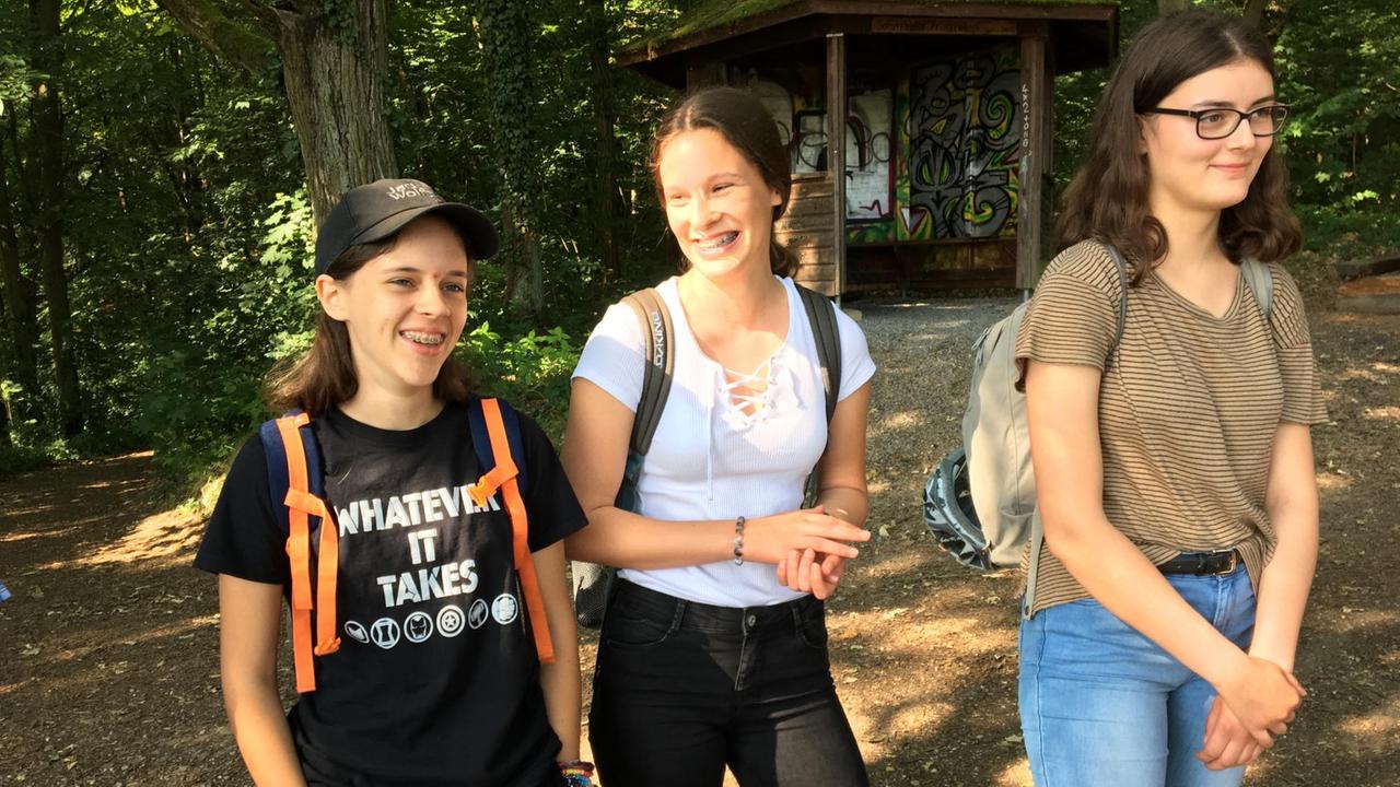Drei junge Frauen gehen über einen Waldweg. Im Hintergrund ist eine Waldhütte zu sehen.