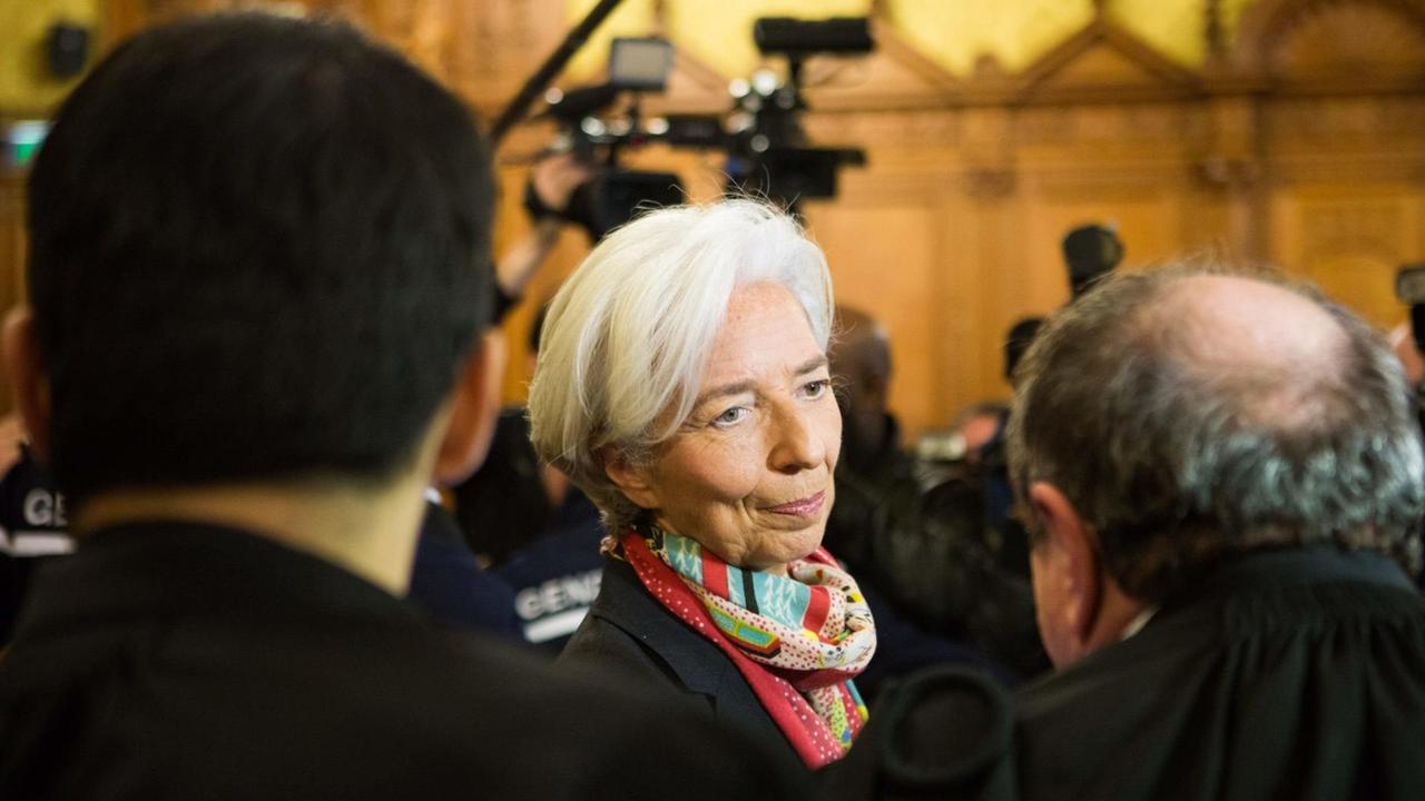Lagarde unterhält sich mit einem Mann, der nur von hinten zu sehen ist; ebenso wie ein zweiter Mann neben ihr. Dahinter sieht man unscharf Pressefotografen, die ihre Kameras in die Höhe recken. Lagarde unterhält sich mit einem Mann, der nur von hinten zu sehen ist; ebenso wie ein zweiter Mann neben ihr. Dahinter sieht man unscharf Pressefotografen, die ihre Kameras in die Höhe recken.