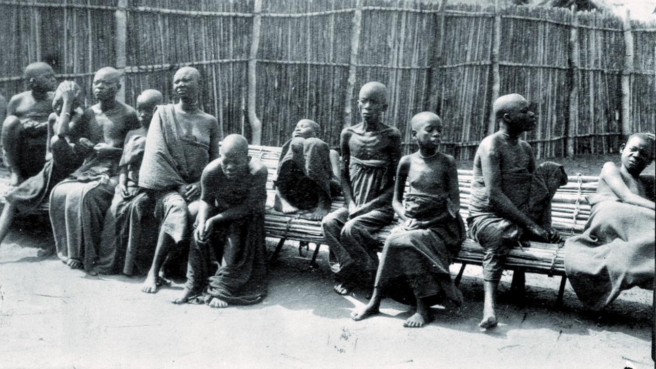 Historisches Foto: Schwarze Patienten mit Tsetse sitzen auf einer Bank. Beim Robert-Koch-Institut ist dieses Foto so betitelt: "Patienten, die an der Schlafkrankheit leiden, dämmern vor sich hin. Dann sterben sie."
Der Begleittext weist allerdings auf die "Schwersten Nebenwirkungen durch Atoxyl" hin und bezeichnet Kochs letzte Forschungsreise gleichzeitig als seine "unrühmlichste". Historisches Foto: Schwarze Patienten mit Tsetse sitzen auf einer Bank. Beim Robert-Koch-Institut ist dieses Foto so betitelt: "Patienten, die an der Schlafkrankheit leiden, dämmern vor sich hin. Dann sterben sie."
Der Begleittext weist allerdings auf die "Schwersten Nebenwirkungen durch Atoxyl" hin und bezeichnet Kochs letzte Forschungsreise gleichzeitig als seine "unrühmlichste".