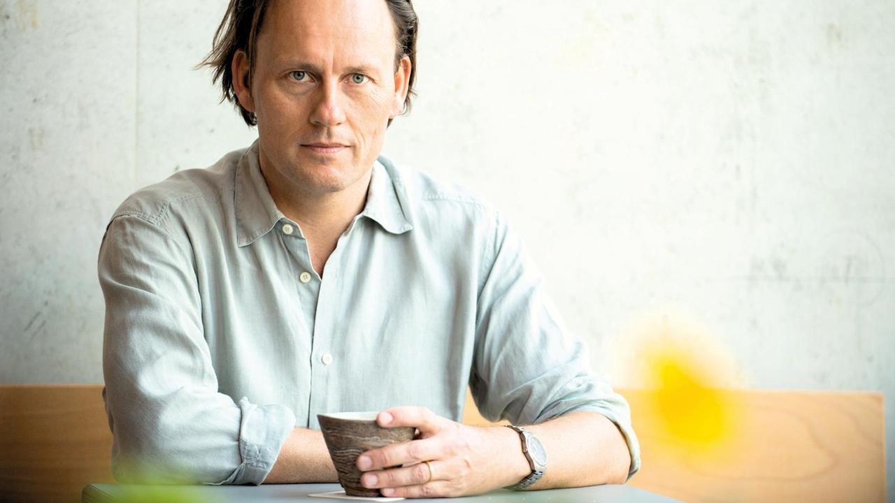Der Autor Gregor Hens sitzt in grauen Hemd vor einer grauen Wand an einem Tisch, in der Hand eine Tasse. Der Autor Gregor Hens sitzt in grauen Hemd vor einer grauen Wand an einem Tisch, in der Hand eine Tasse.