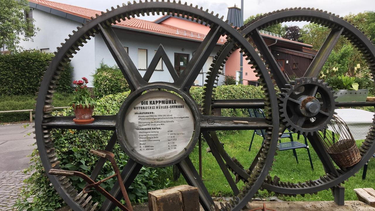 Zwei Zahnräder im Hof der Kappmühle im Dorf Mackenzell in der hessischen Rhön mit der Aufschrift "Historisches Mühlengetriebe"