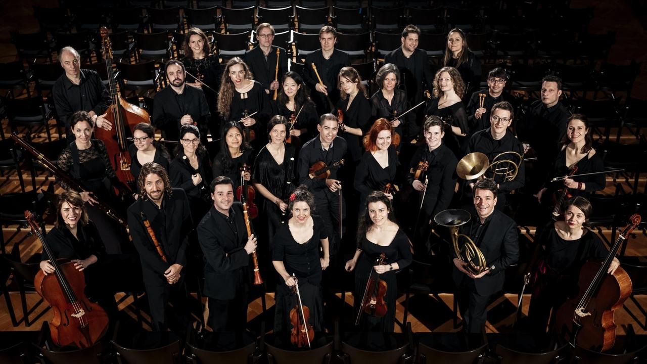 Eine Gruppe von Musikern steht dicht gedrängt in einem schlichten Zuschauerraum mit schwarzen, modernen Stühlen und schaut nach oben in die Kamera. Eine Gruppe von Musikern steht dicht gedrängt in einem schlichten Zuschauerraum mit schwarzen, modernen Stühlen und schaut nach oben in die Kamera.