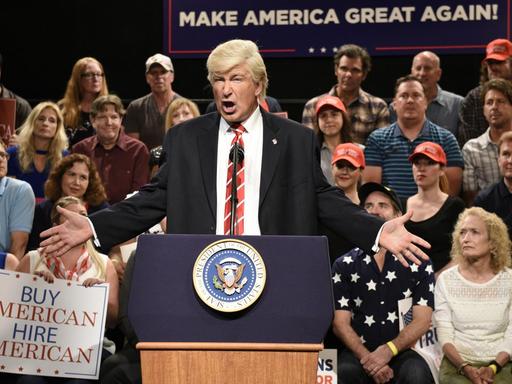 Alec Baldwin steht in seiner Rolle als Donald Trump verkleidet an einem Redepult.