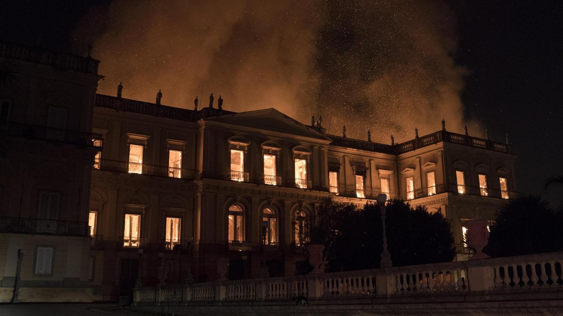 Das Bild zeigt das brennende Gebäude in der Nacht, durch die Fenster sieht man die Flammen, Rauch und Funken steigen in den Nachthimmel.