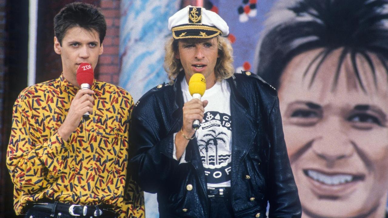 Ein Foto von 1989 von Günther Jauch und Thomas Gottschalk: Sie moderieren das ZDF Mittagsmagazin. Jauch trägt dabei ein bund gemustertes Hemd, Gottschalk eine Lederjacke und Kapitänsmütze.