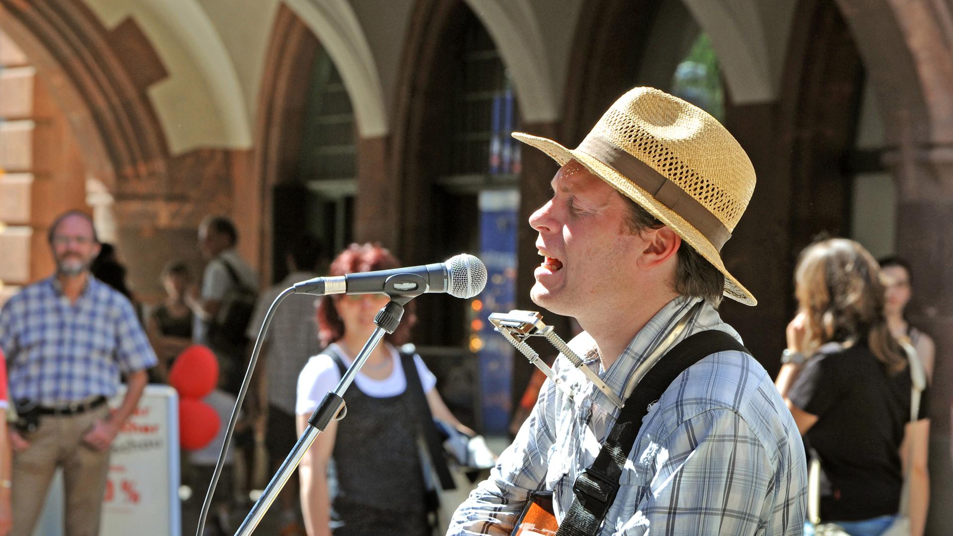 Der Musiker Jimmy Kelly singt im Stadtzentrum von Leipzig als Straßenmusikant. Er trägt einen Strohsonnenhut und spielt Gitarre.