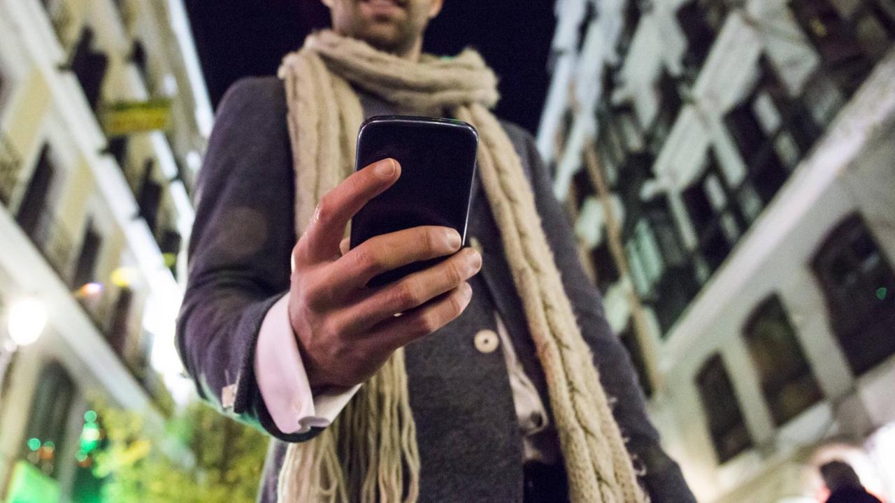 Ein Mann in einer bei Dunkelheit beleuchteten Fußgängerzone hält ein Smartphone in der Hand.