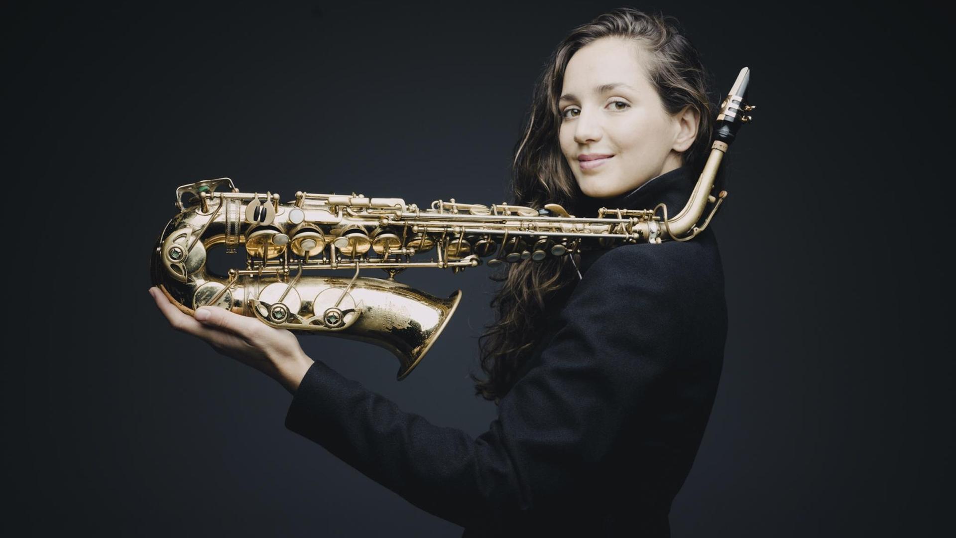 Eine Frau mit langen braunen Haaren hält ein Saxofon in der Hand. Sie balanciert es auf der linken Schulter und der ausgetreckten linken Hand. Es ist die Saxofonistin Asya Fateyeva. Eine Frau mit langen braunen Haaren hält ein Saxofon in der Hand. Sie balanciert es auf der linken Schulter und der ausgetreckten linken Hand. Es ist die Saxofonistin Asya Fateyeva.