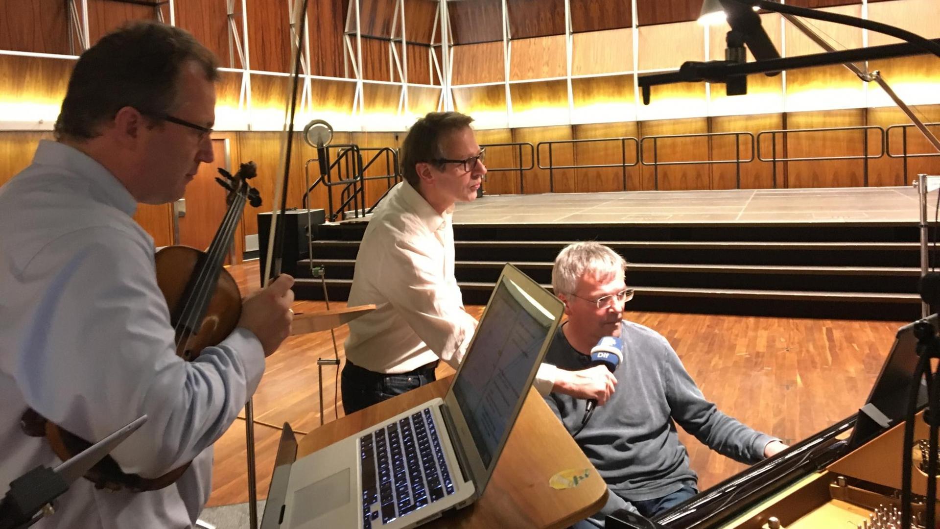 Am Flügel sitzt der Pianist und schaut in die Noten auf dem Notenpult, ebenso der Moderator und der Geiger links danebenstehend, vor dem ein Laptop auf einem Notenständer steht.. Am Flügel sitzt der Pianist und schaut in die Noten auf dem Notenpult, ebenso der Moderator und der Geiger links danebenstehend, vor dem ein Laptop auf einem Notenständer steht..