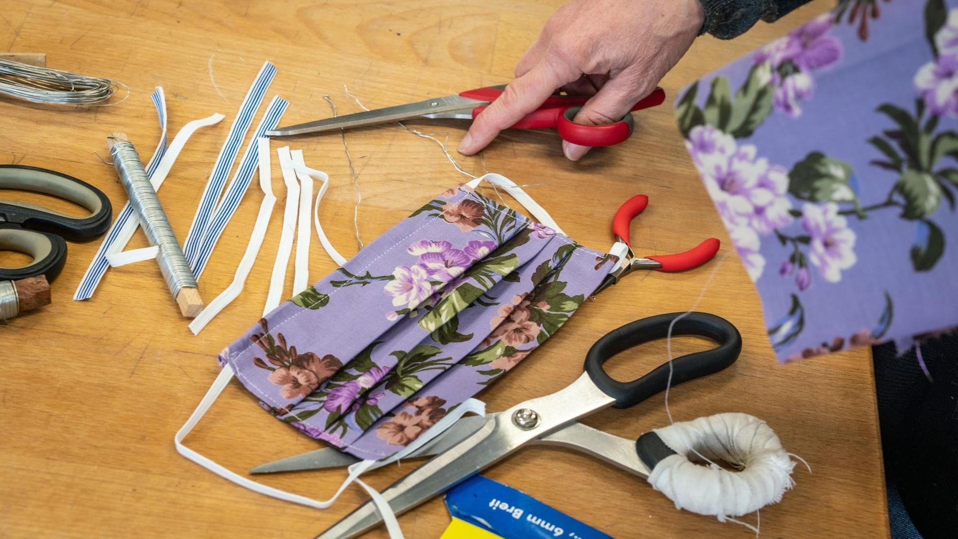 Auf einem Tisch liegt ein fertig genähter Mundschutz mit floralem Muster auf lila Hintergrund sowie diverse Utensilien wie Scheren, Zange, Gummibänder und eine Rolle Draht. Auf einem Tisch liegt ein fertig genähter Mundschutz mit floralem Muster auf lila Hintergrund sowie diverse Utensilien wie Scheren, Zange, Gummibänder und eine Rolle Draht.