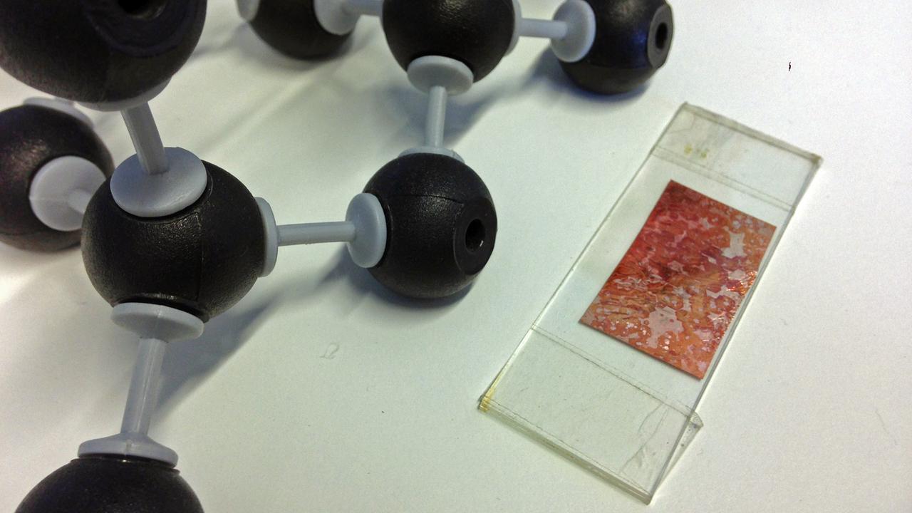 Eine Kupferfolie auf der Graphen gewachsen ist liegt neben einem Atommodell. Eine Kupferfolie auf der Graphen gewachsen ist liegt neben einem Atommodell.