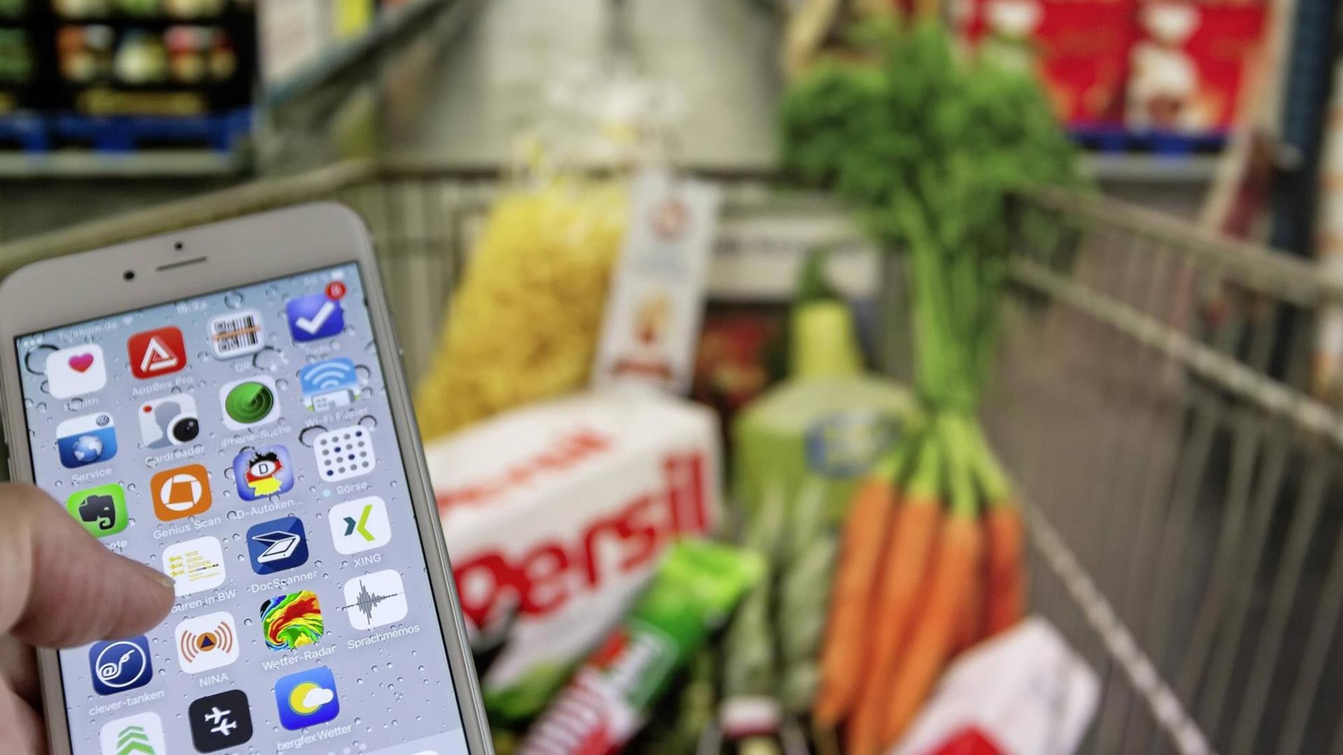 Ein Smarthphone mit der NINA-Warn-App vor einem gefüllten Einkaufswagen in einem Supermarkt
