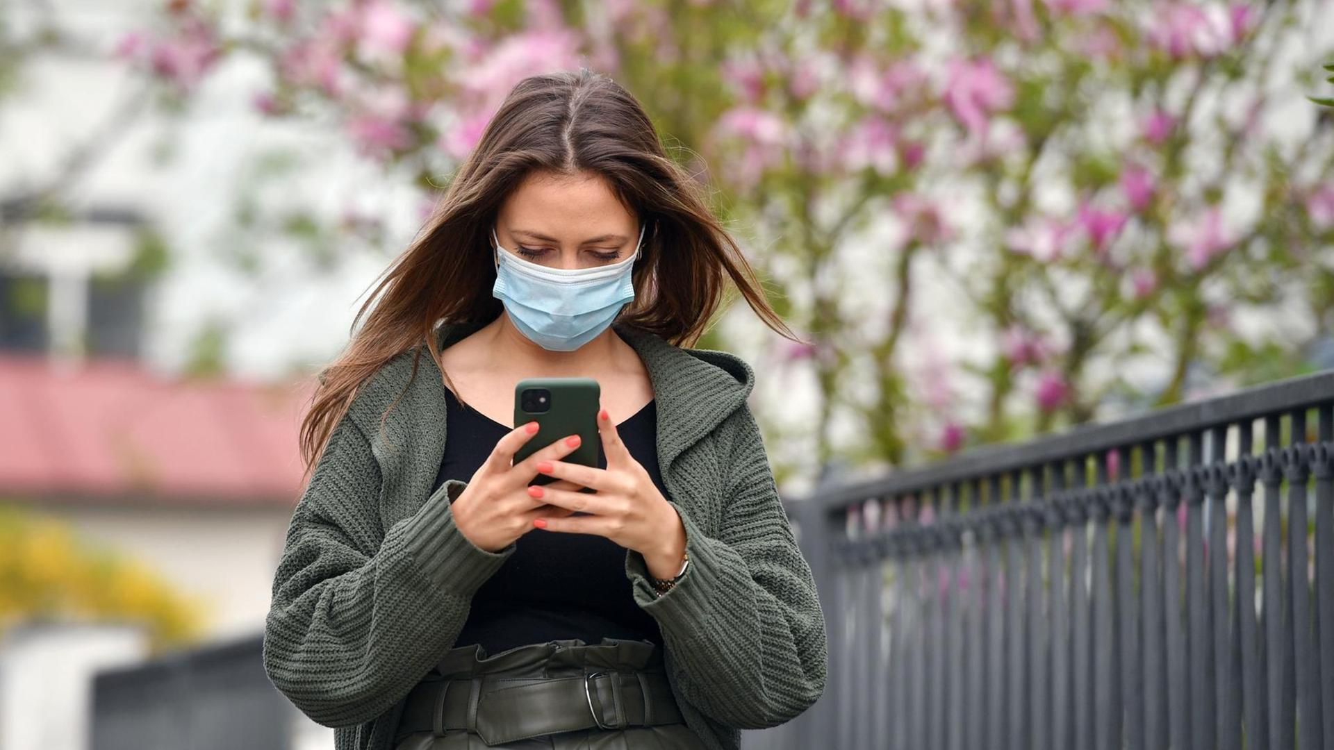 Eine junge Frau mit Schutzmaske blickt auf ihr Smartphone. (Symbolbild) Eine junge Frau mit Schutzmaske blickt auf ihr Smartphone. (Symbolbild)