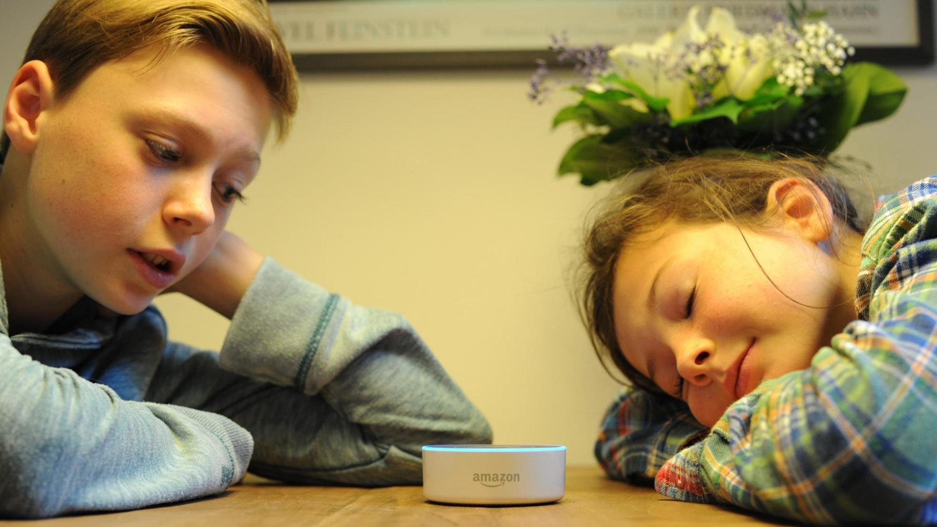 Ein Junge spricht in den Lautsprecher Echo Dot von Amazon, ein Mädchen mit geschlossenen Augen ihm gegenüber hat den Kopf auf den Tisch gelegt. Ein Junge spricht in den Lautsprecher Echo Dot von Amazon, ein Mädchen mit geschlossenen Augen ihm gegenüber hat den Kopf auf den Tisch gelegt.
