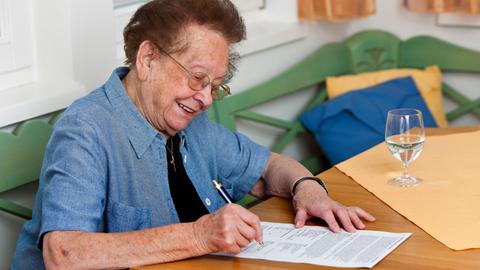 Eine ältere brünette Frau mit schwarzem T-Shirt und blauer Jacke darüber sitzt auf einer grünen Bank an einem Tisch und unterschreibt einen Vertrag. Auf dem Tisch steht ein Glas Wasser.