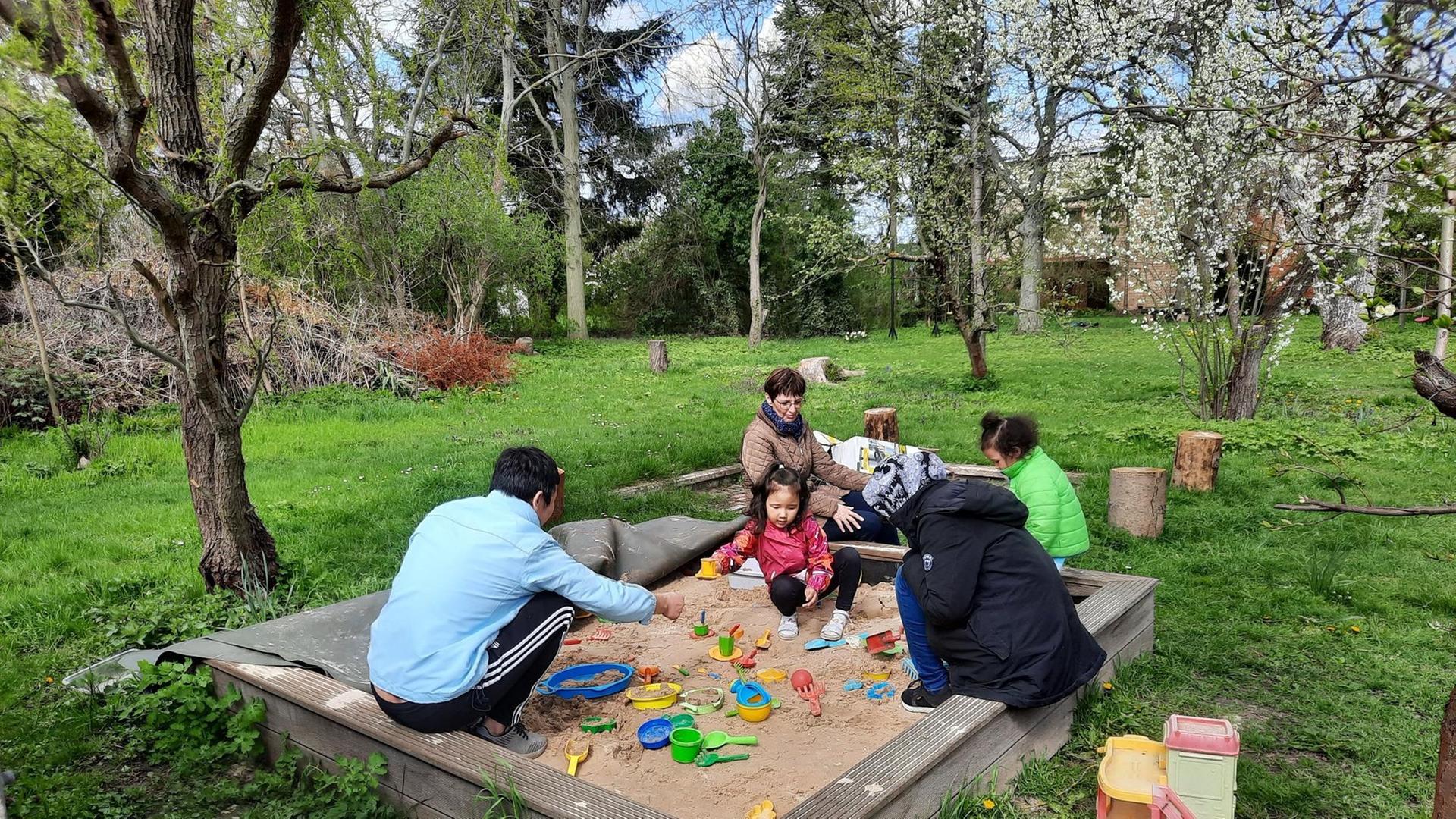 In einem großen Garten mit zahlreichen Bäumen steht ein Sandkasten, in dem zwei kleine dunkelhaarige Mädchen spielen. Auf dem Rand sitzen die Eltern und eine weitere Frau. In einem großen Garten mit zahlreichen Bäumen steht ein Sandkasten, in dem zwei kleine dunkelhaarige Mädchen spielen. Auf dem Rand sitzen die Eltern und eine weitere Frau.