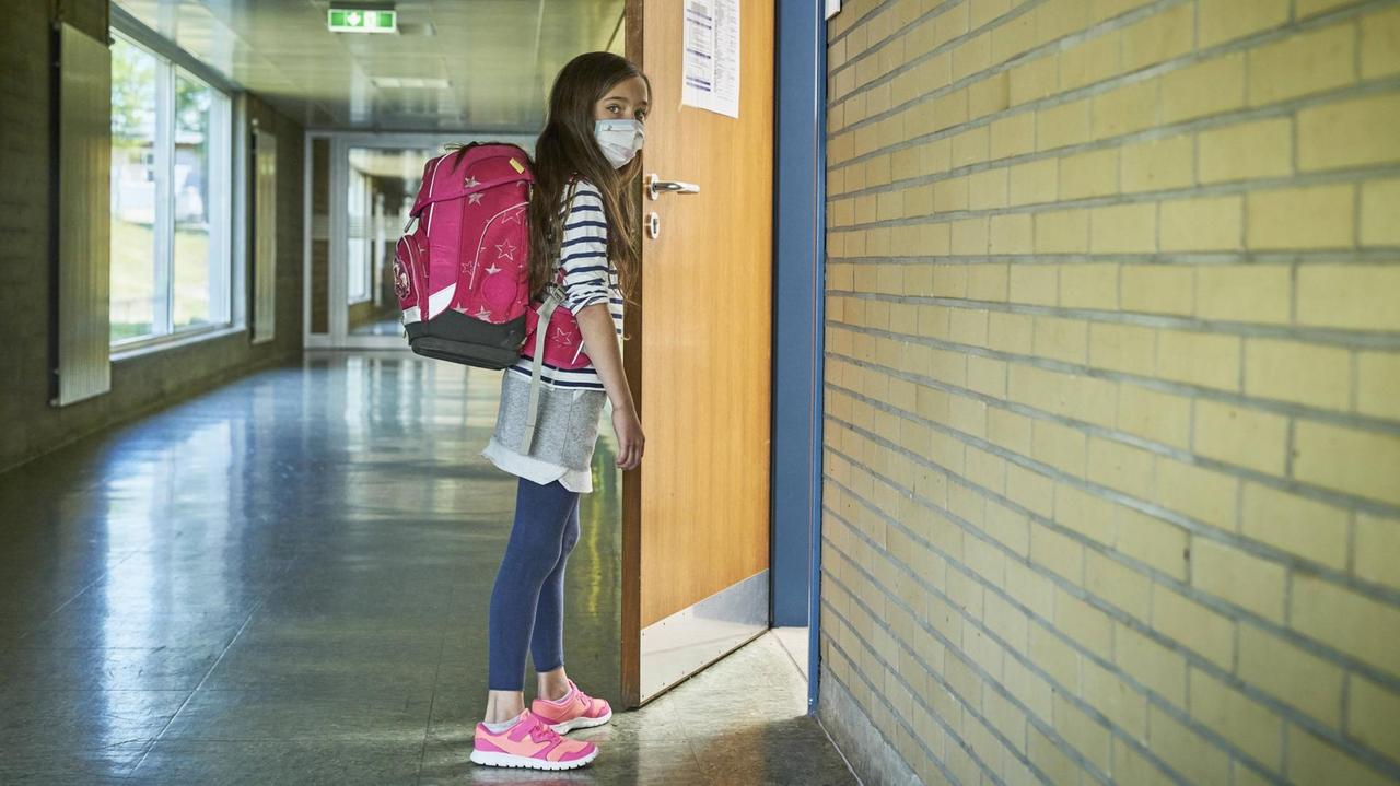 Ein Mädchen mit Mund-Nasen-Maske öffnet die Tür zu einem Klassenraum. Ein Mädchen mit Mund-Nasen-Maske öffnet die Tür zu einem Klassenraum.