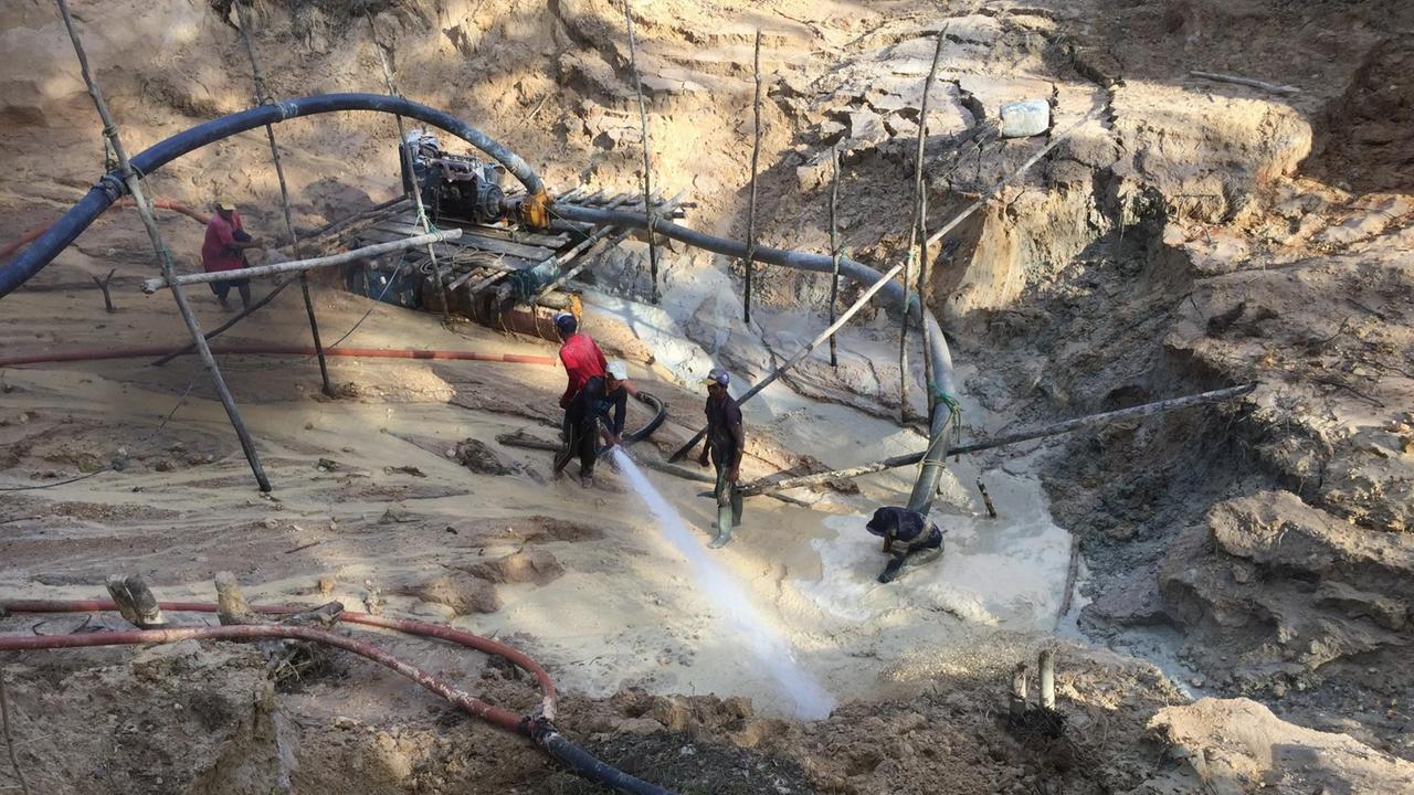 Goldgräber stehen am Abhang im Schlamm und spritzen mit einer Pumpe Wasser ins Gestein. Goldgräber stehen am Abhang im Schlamm und spritzen mit einer Pumpe Wasser ins Gestein.